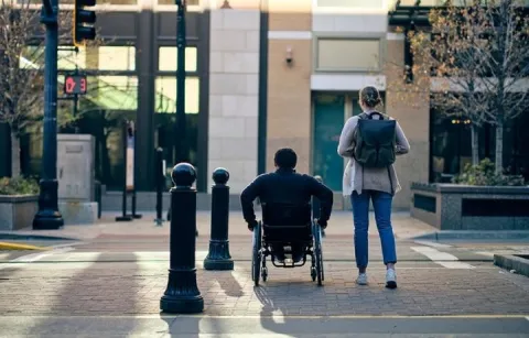 A person in wheelchair and another standing person at a city sidewalk